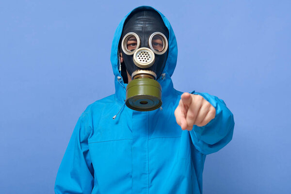 Closeup portrait of ecologist frights with factories which pollute air, water, forests and land, scientist wearing uniform and respirator, pointing to camera with with fore finger against blue wall.