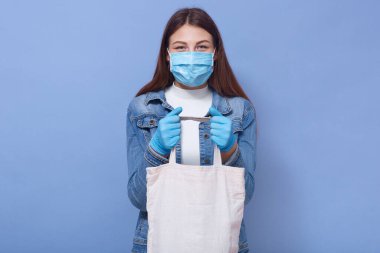 Kameraya doğrudan bakan dikkatli genç bir kadının yatay stüdyo fotoğrafı. Elinde alışveriş torbası, koruyucu maske ve eldiven var. Karantina sırasında alışveriş yapıyor. Covid 19 konsepti.