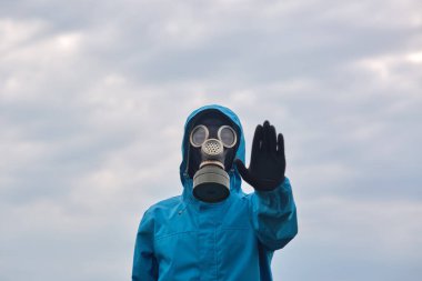 Bir el kaldıran, gaz maskesi takan, eldiven takan ve koruma elbisesi giyen, özel bir göreve katılan, gökyüzü arka planında poz veren bir adamın yatay dış görüntüsü. Halk ve felaket konsepti.