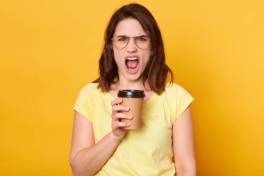Stüdyoda, sarı stüdyo arkasında izole edilmiş, kahve fincanı tutan, doğrudan kameraya bakan, bir şeyler bağıran, sıradan bir tişört giyen kızgın bir kadının görüntüsü. İnsan duyguları kavramı.