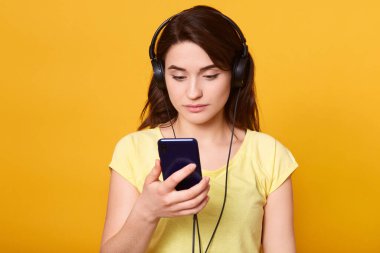 Koyu renk tişört giyen, modern akıllı telefonu elinde tutan, kulaklıkla poz veren, favori şarkıları olan müzik listesini seçen koyu renk saçlı kadın portresi müzik dinlemeyi seviyor..