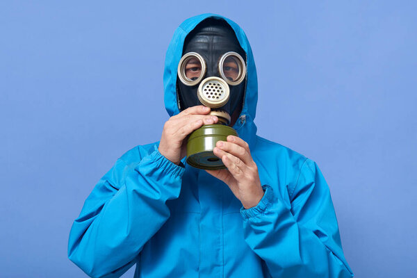 Portrait of liquidating agent touching his gas mask, regulating, showing way of usage, being instructor, wearing uniform, posing isolated over blue background in studio. Environmental problems.