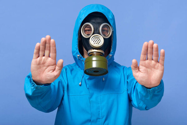Image of careful man making gesture, raising arms, keeping distance, wearing blue jacket with hood, posing isolated over blue background, ecological trouble, having any fresh air to breathe.