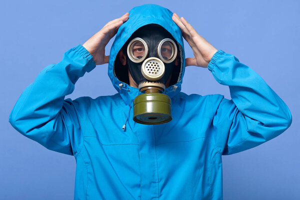 Horizontal portrait of attentive guy putting on hood, wearing gas mask and blue jacket, raising hands, looking directly at camera, being in difficult dangerous situation. Liquidating agent concept.