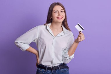 Bir elinde kredi kartı tutan komik, çekici genç bir kadının portresi, samimi bir şekilde gülümsüyor, doğrudan kameraya bakıyor, kot pantolon ve açık renk gömlek giyiyor, elini belinde tutuyor. Gider kavramı.