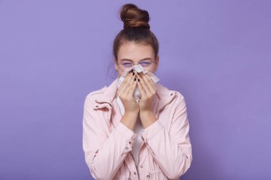 Hasta genç bayanın burnunu sildiği, mendil kullandığı, burnu aktığı, kendini kötü hissettiği, günlük kıyafetler giydiği, eflatun arka planda yalnız poz verdiği kapalı bir çekim. Sağlık sorunları kavramı.