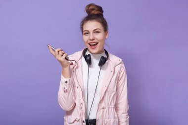 Mutlu, heyecanlı, günlük kıyafetler giyen, kulaklık takan, modern akıllı telefonu elinde tutan, ağzı açık bir şekilde leylak duvara poz veren, müzik dinlemekten hoşlanan güzel bir kadın..