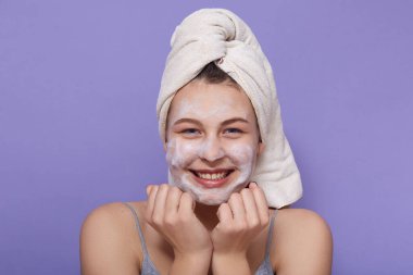Neşe dolu, güzel görünümlü, ıslak saçları yuvarlanan, ellerini yüzüne yakın tutan, spa yapan, stüdyodaki leylak arkaplan üzerinde izole edilmiş poz veren genç bayanın yakın portresi. Güzellik konsepti.