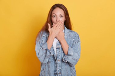 Etkilenmiş karizmatik genç modelin yatay görüntüsü geniş çapta gözleri açıyor, elleri ağızla örtüyor, stüdyoda sarı arka planda tek başına duruyor, doğrudan kameraya bakıyor. Duygular konsepti.