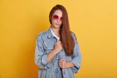 Uzun saçlı, doğrudan kameraya bakan, güneş gözlüğü ve kot ceket giyen, sarı arka planda izole edilmiş poz veren güzel, tatlı, narin bir genç kızın yatay fotoğrafı..