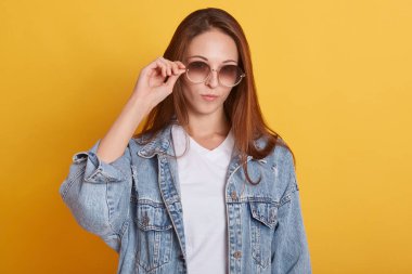 Stüdyoda sarı arka planda izole edilmiş, moda güneş gözlüğü ve kot ceket giyen, yalnız olan, dinlenen çekici bir gencin yakın portresi. Gençlik ve tarz kavramı.