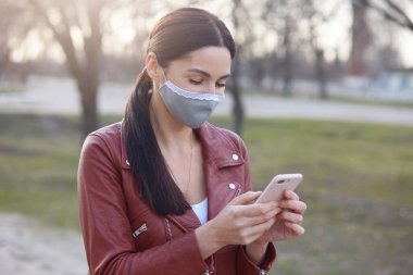 Açık havada, akıllı telefon tutan, cihaz kullanan, koruyucu maske takan, sosyal ağ sitelerini kontrol eden parkta yürüyen, çevrimiçi iletişim kuran esmer bir kızın görüntüsü. Covid19 konsepti.