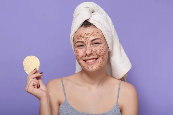 Attractive young woman with her hair wrapped in white towel, girl with perfect skin cleansing her face, using coffee scrub, posing isolated over lilac background, holding cotton sponge in hand.