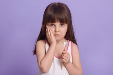 Zavallı üzgün çocuğun yatay portresi, hayal kırıklığına uğramış yüz ifadesi, üzgün olmak, diş ağrısı, diş fırçası tutmak, doğrudan kameraya bakmak, uyku gömleği giymek. Çocuk konsepti.