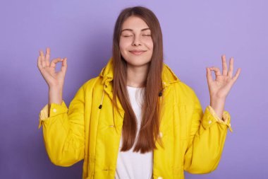 Kapalı gözlerle gülümseyen mutlu bir kadının yakın plan portresi, ön parmaklarını ve başparmağını bir arada tutan, sarı ceket giyen, koyu renk saçlı, eflatun rengi duvarda duran, rahatlatıcı bir kız..