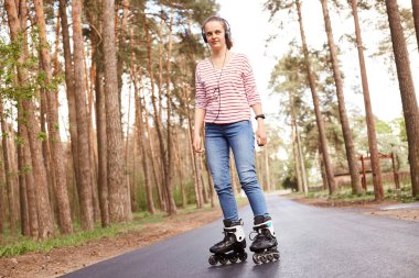 Keyifli genç kadın paten kayarken, orman yolunda tek başına müzik dinlerken, dinlenirken, aktivitelere düşkün olurken, günlük giysiler giyerken. Dinlenme kavramı.