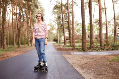 At kuyruklu, günlük kıyafetler giyen, ormanda paten kayarken kulaklıkla müzik dinleyen, dışarıda vakit geçiren, asfalt yolda paten kayan güzel bir kız..