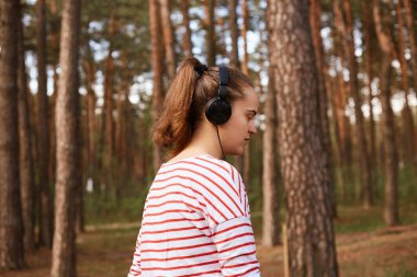 At kuyruklu, çizgili bir tişört giyen, kulaklıkla müzik dinleyen, açık havada dinlenen, ağaçlarla çevrili ağaçlarla çevrili bir şekilde fotoğraflanmış bir kızın yan görüntüsü. İnsanlar.