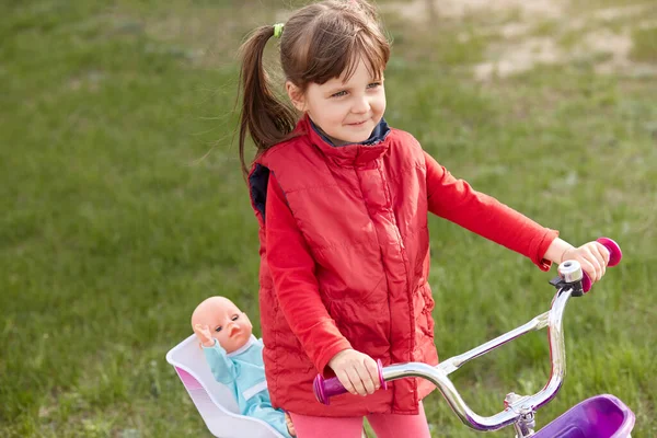 子供の座席で彼女の人形と自転車に乗る小さな魅力的な女の子の屋外ショットは 女性の子供が背景に緑の芝生と魅力的な笑顔でポーズをオープンエアで時間を過ごす 幼少期の概念 — ストック写真