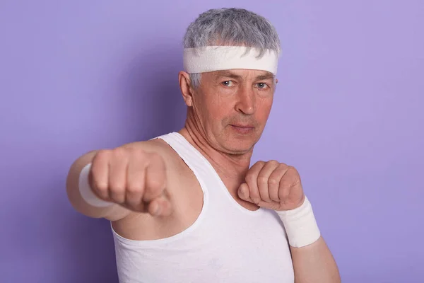 Studio picture of sporty confident man looking directly at camera, showing his fists, wearing white shirt and headband, having box training, learning punch, being concentrated. Boxing concept.