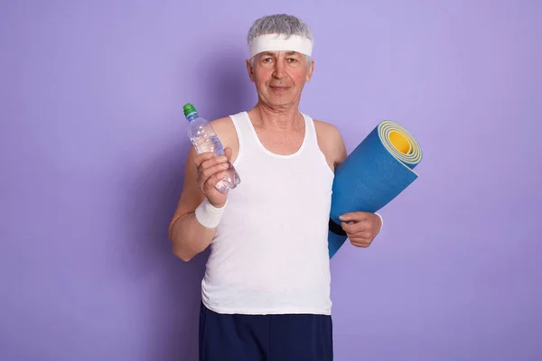 Indoor portrait of energetic happy senior man looking directly at camera, holding rug and bottle of water in both hands, keeping fit, wearing headband, shirt and shorts. People and sport concept.