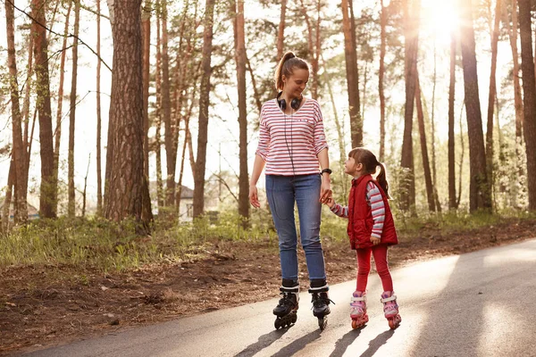 Outdoor shot of mother with her daughter spending time together in forest, enjoying to rollerblade outdoor, being happy, expressing joy, being in good mood, looking at each other happily. Lifestyle.