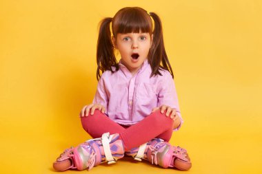 Şaşkınlık içindeki sevimli kız çocuğunun görüntüsü, sarı stüdyo arka planında izole edilmiş, ağzı açık poz veren, şok olan, bacak bacak bacak üstüne atmış paten kayan küçük kız. Çocuğun duyguları.