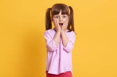 Şok olmuş ya da şaşırmış, ağzı açık poz veren, ellerini yanaklarından ayırmayan, hayrete düşmüş yüz ifadeleriyle doğrudan kameraya bakan güzel kızın yakın portresi. Çocukluk.