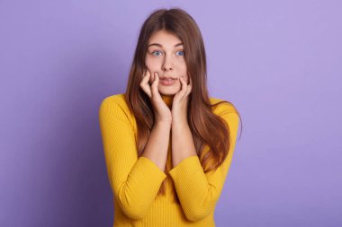 Uzun saçlı, korkmuş bir kadın, kameraya büyük gözlü, bir şeyden korkan, sarı günlük gömlek giyen, leylak rengi stüdyo arka planında yalnız poz veren bir kız.