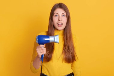 Elinde mavi saç kurutma makinesi olan güzel bir kadının yatay görüntüsü, sarı stüdyo arka planında izole edilmiş poz veriyor, şaşkın bir yüz ifadesi var, şaşırmış görünüyor, ağzı açık poz veriyor.