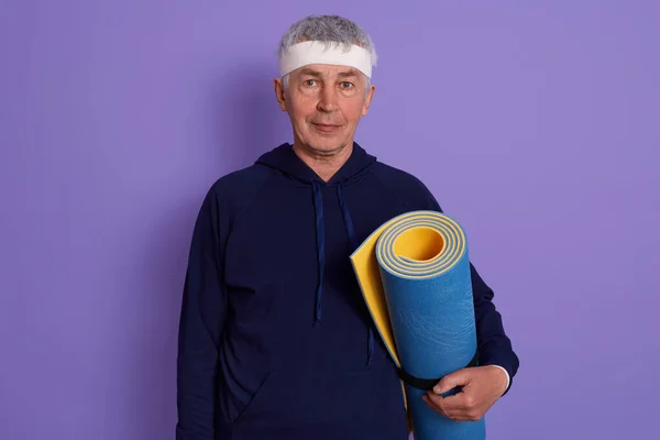 Horizontal shot of senior white haired male with head band and holding blue yoga mat in hands, posing isolated over lilac background, being ready to do physical exercises. Active old age concept.