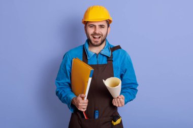 Stüdyoda mavi arka planda tek başına duran, elinde kağıtlar olan, ağzı açık, bağıran, sakallı, mavi önlüklü, kasklı ve kahverengi önlüklü duygusal yakışıklı bir müteahhit portresi..
