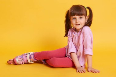 Stüdyoda, iki at kuyruklu, doğrudan kameraya bakan, stüdyoda sarı arka planda izole edilmiş, tekerlekli paten giyen şirin bir çocuk fotoğrafı. Çocukluk oyunları konsepti.