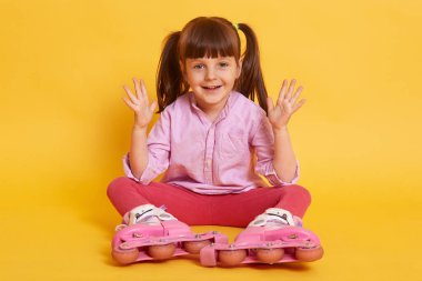 Oyuncu, enerjik, elini kaldıran, sarı arka planda izole bir şekilde yerde oturan pembe günlük giysiler ve tekerlekli paten giyen, samimi bir şekilde gülümseyen küçük kızın kapalı alanda çekilmiş resmi. Eğlenceli konsept.