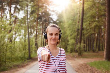 Sevimli, güzel, pozitif görünümlü genç bir bayanın yakından çekilmiş portresi samimi bir şekilde gülümsüyor, iyi bir ruh hali içinde, işaret parmağıyla kameraya bakıyor, doğrudan kameraya bakıyor, müzik dinliyor. Dinlenme kavramı.