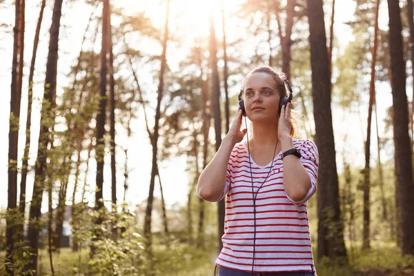 ヘッドフォンを頭の上に持ち 音楽に耳を傾け 平和的な表情を持ち 先を見て 周りの自然を楽しんでいる優しい良い若い女性の外観の肖像画の外 休憩の概念 — ストック写真