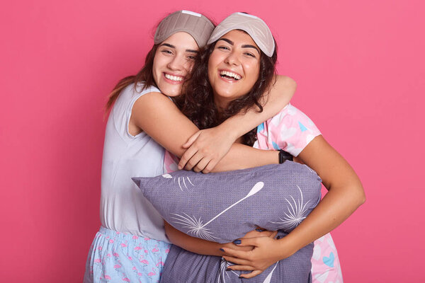 Улыбающаяся молодая белая женщина обнимает свою подругу с подушкой в руке на розовом фоне. Красивые девушки в пижамах и спальных складках стоят вместе и улыбаются
.
