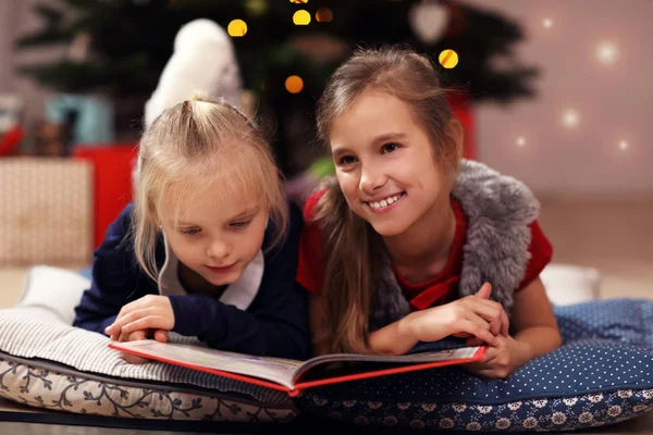 Duas irmãs pequenas bonitos lendo livro de história juntos sob a árvore de Natal — Fotografia de Stock