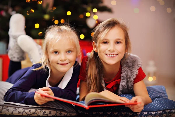 Dos monas hermanitas leyendo el libro de cuentos juntas bajo el árbol de Navidad —  Fotos de Stock