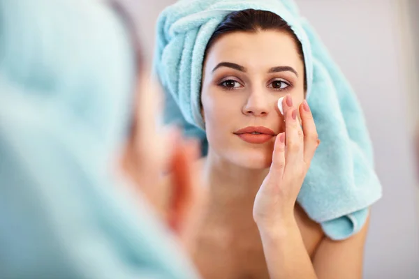 Jovem mulher de pé no banheiro e aplicando creme facial pela manhã — Fotografia de Stock