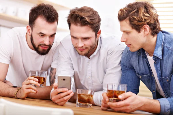 Amigos alegres que se divierten con el teléfono inteligente y beber en casa — Foto de Stock