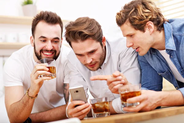 Amigos alegres se divertindo com smartphone e bebendo em casa — Fotografia de Stock