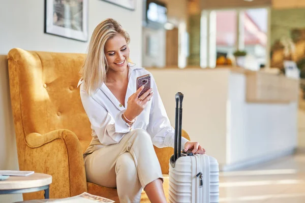 Businesswoman with luggage in modern hotel lobby using smartphone — Stock Photo, Image