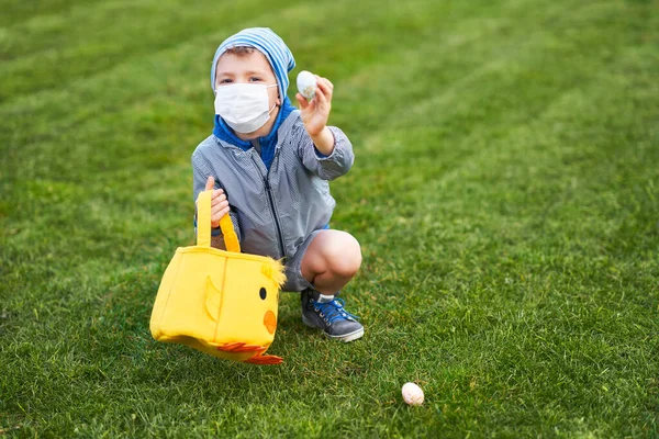 Liten pojke bär skyddsmask jakt på påskägg i vår trädgård. — Stockfoto