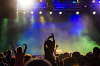 Konser kalabalık bir konser, insan siluetleri katılıyor mu görünür, arkadan aydınlatmalı sahne ışıkları tarafından. Yükseltilmiş elleri ve akıllı telefonlar orada burada görülebilir.