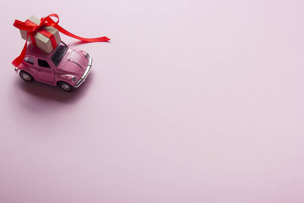 Розовый ретро игрушечный автомобиль, доставляющий подарочную коробку на пастельном фоне. Вид сверху
