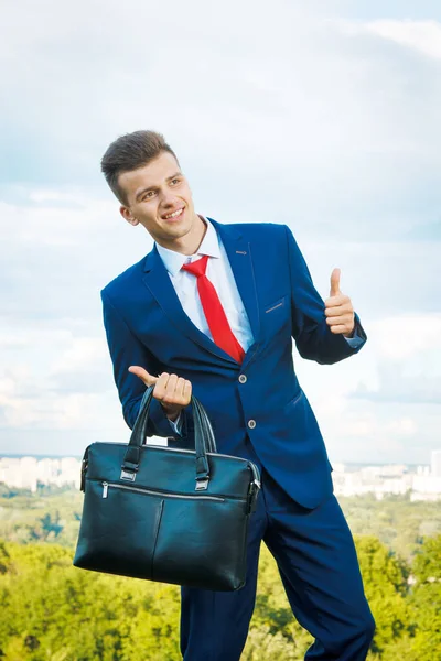 Cheerful smiling businessman who made a good deal dressed in blue suit and red tie with a briefcase in his hand against the backdrop of the city and the sky