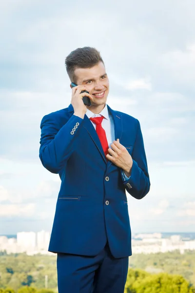 都市と空を背景に彼の手のブリーフケースと青いスーツと赤いネクタイを身に着けている良い取引をした陽気な笑顔のビジネスマン — ストック写真