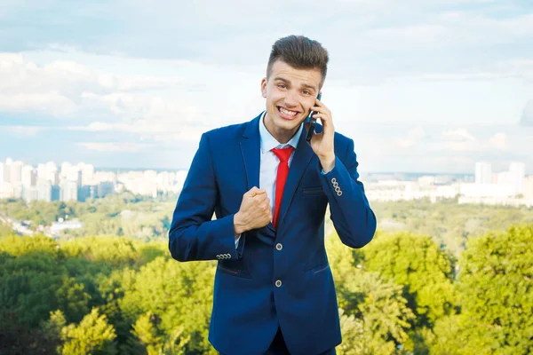 Cheerful smiling businessman who made a good deal dressed in blue suit and red tie with a briefcase in his hand against the backdrop of the city and the sky