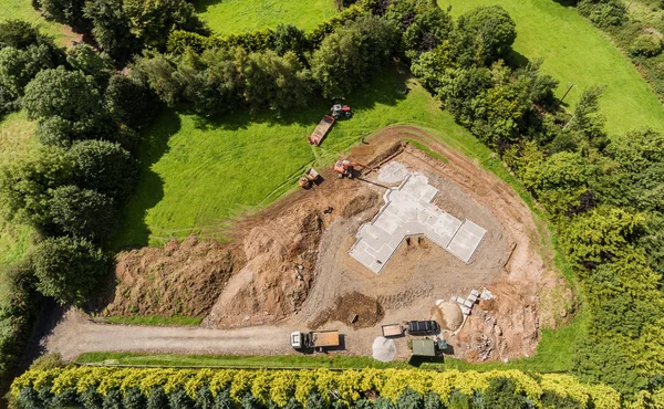 Εναέρια πτηνών μάτι θέα ενός νέου ιδρύματος σκυροδέματος κατασκευή για ένα σπίτι στην εξοχή. Πράσινο χωράφι με μηχανήματα digger και ανατρεπόμενο φορτηγό και τρακτέρ. — Φωτογραφία Αρχείου
